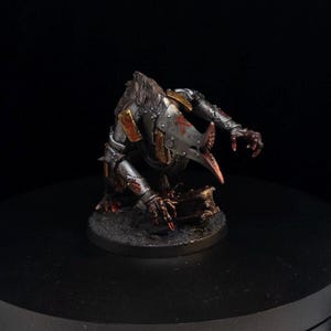 Può includere: Figurina in miniatura di una creatura con becco simile a un uccello e mani artigliate. La figura è dipinta in argento metallizzato, oro e rosso, con un aspetto dettagliato e strutturato. La creatura è posizionata su una base circolare.
