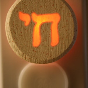 May include: A white ceramic nightlight with a round shape and a cutout of the Hebrew letter "Chai" in orange. The nightlight is plugged into an electrical outlet.