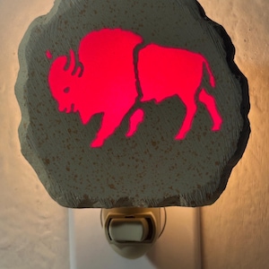 May include: A night light featuring a red bison silhouette on a gray, stone-shaped base. The night light plugs into a standard wall outlet and emits a soft, warm glow. The bison design is a prominent feature.