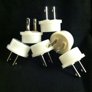 May include: A group of six white electrical plug adapters with two prongs each. The adapters are arranged in a pile on a black surface.
