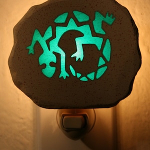 May include: A white ceramic night light shaped like a rock with a green lizard silhouette cutout. The night light is plugged into a standard electrical outlet.