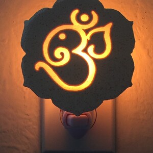 May include: A decorative night light featuring the Om symbol, illuminated with a warm glow. The light is encased in a gray, petal-shaped frame and plugs into a standard wall outlet. The Om symbol is a spiritual icon.