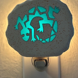 May include: A night light featuring a teal lizard design on a stone-like surface. The lizard glows when the light is on, creating a soft, ambient glow. The night light plugs directly into a standard wall outlet.