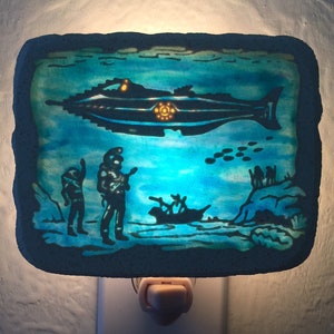 May include: A blue nightlight with a scene of a submarine, two figures, and a small boat in the ocean. The nightlight is made of a textured material and has a white electrical cord.