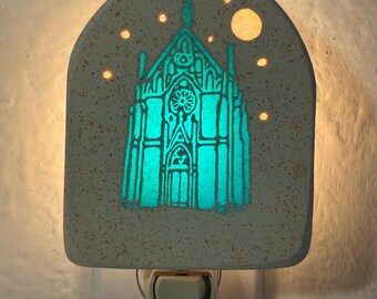 May include: A decorative night light featuring a church design with a glowing turquoise interior. The light is encased in a speckled gray stone-like material, with a crescent top and a plug-in base. The design includes a church with a rose window and stars.