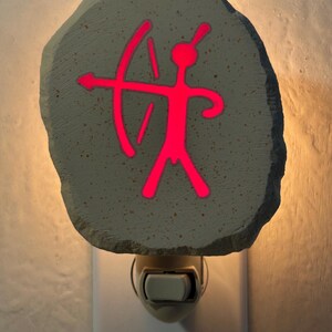 May include: A decorative night light featuring a gray stone-like face with a glowing red archer figure. The night light plugs into a standard wall outlet and has an on/off switch. The design is reminiscent of ancient cave art.