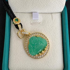 May include: A gold pendant featuring a carved green jade-colored Buddha figure surrounded by a circular frame of small clear stones. The pendant hangs from a black cord with a gold accent piece containing a small green stone and clear crystals.