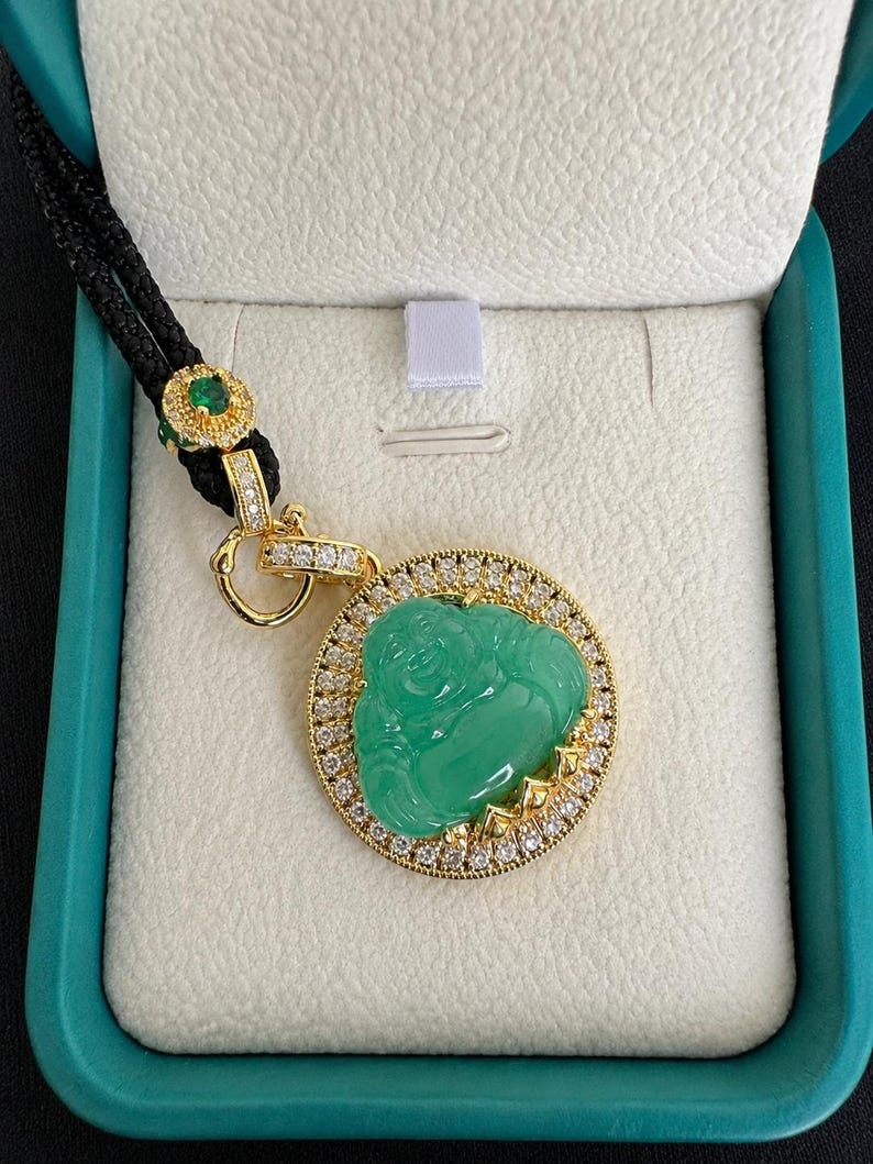 May include: A gold pendant featuring a carved green jade-colored Buddha figure surrounded by a circular frame of small clear stones. The pendant hangs from a black cord with a gold accent piece containing a small green stone and clear crystals.