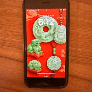 May include: A smartphone displays a red background with jade pendants. The pendants include a dragon, a laughing Buddha, a yin-yang symbol, and two small lion statues. The time is 6:12 AM on Monday, July 7th. The phone is black.