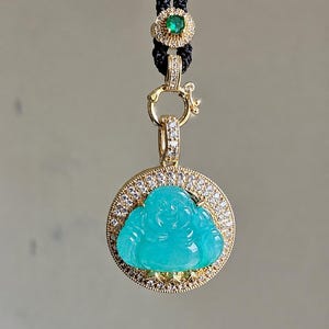May include: A pendant featuring a carved turquoise-colored Buddha figure set within a circular gold frame encrusted with small white diamonds. The pendant hangs from a black cord adorned with a small gold accent piece featuring a green gemstone.