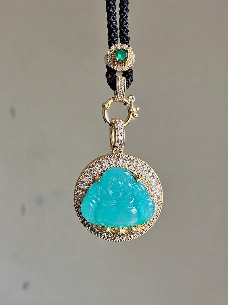 May include: A pendant featuring a carved turquoise-colored Buddha figure set within a circular gold frame encrusted with small white diamonds. The pendant hangs from a black cord adorned with a small gold accent piece featuring a green gemstone.