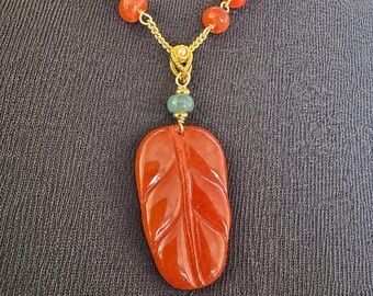 May include: A necklace featuring a carved orange leaf-shaped pendant suspended from a gold-tone chain. The chain is accented with round orange beads and a single light green bead positioned directly above the leaf pendant.