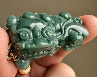 May include: A carved green jade pendant shaped like a mythical creature with prominent eyes, a wide snout, and detailed facial features. The pendant is attached to a cord featuring small round beads in shades of green, yellow, and red with gold accents.