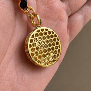 May include: A gold-toned circular pendant featuring a raised honeycomb pattern on its surface. The pendant is attached to a black cord by a gold-toned jump ring and a decorative gold-toned connector piece with small black accents.