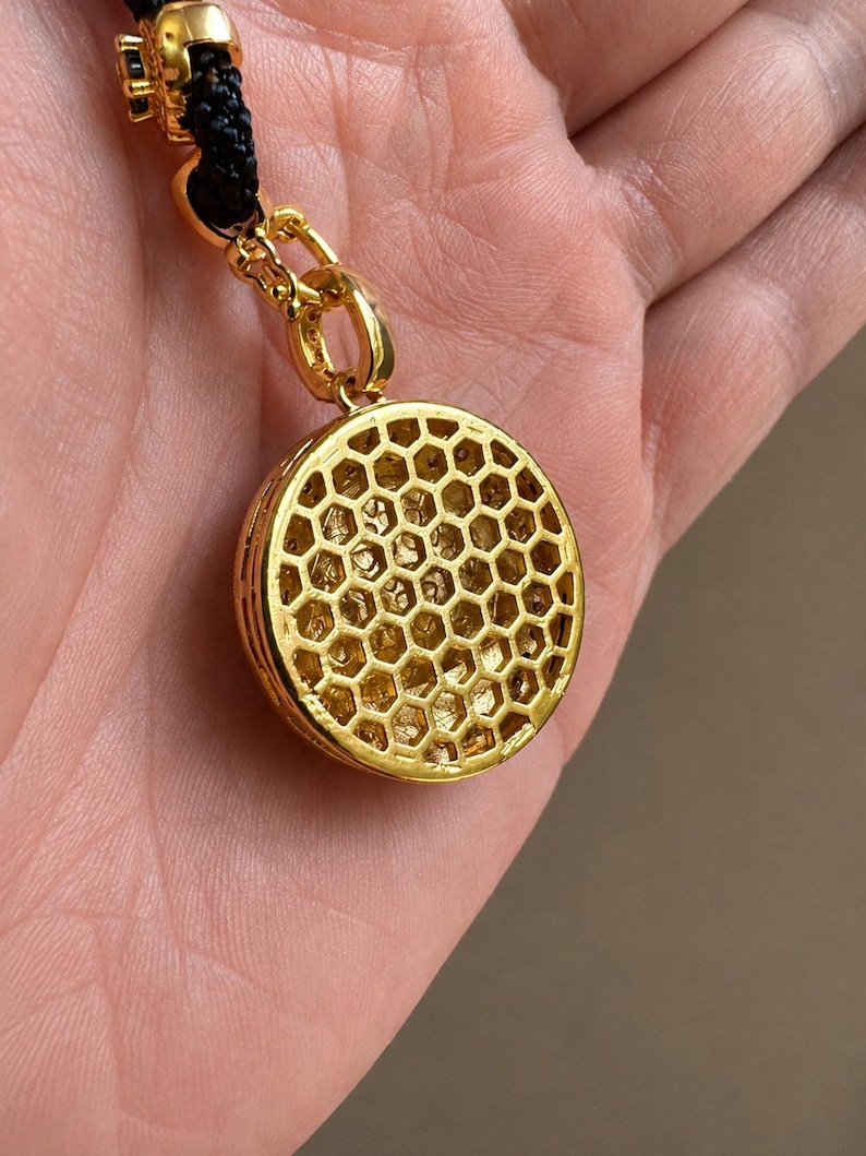 May include: A gold-toned circular pendant featuring a raised honeycomb pattern on its surface. The pendant is attached to a black cord by a gold-toned jump ring and a decorative gold-toned connector piece with small black accents.
