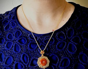 Puede incluir: Un collar con colgante dorado con una piedra circular de color marrón rojizo engastada en un marco de filigrana ornamentado. El collar se luce sobre una blusa azul oscuro texturizada.