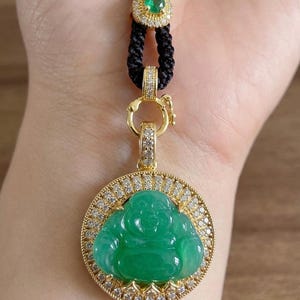 May include: A circular pendant featuring a carved green jade Buddha figure set in a gold frame encrusted with small white diamonds. The pendant hangs from a black braided cord adorned with a small gold accent piece containing a green stone.