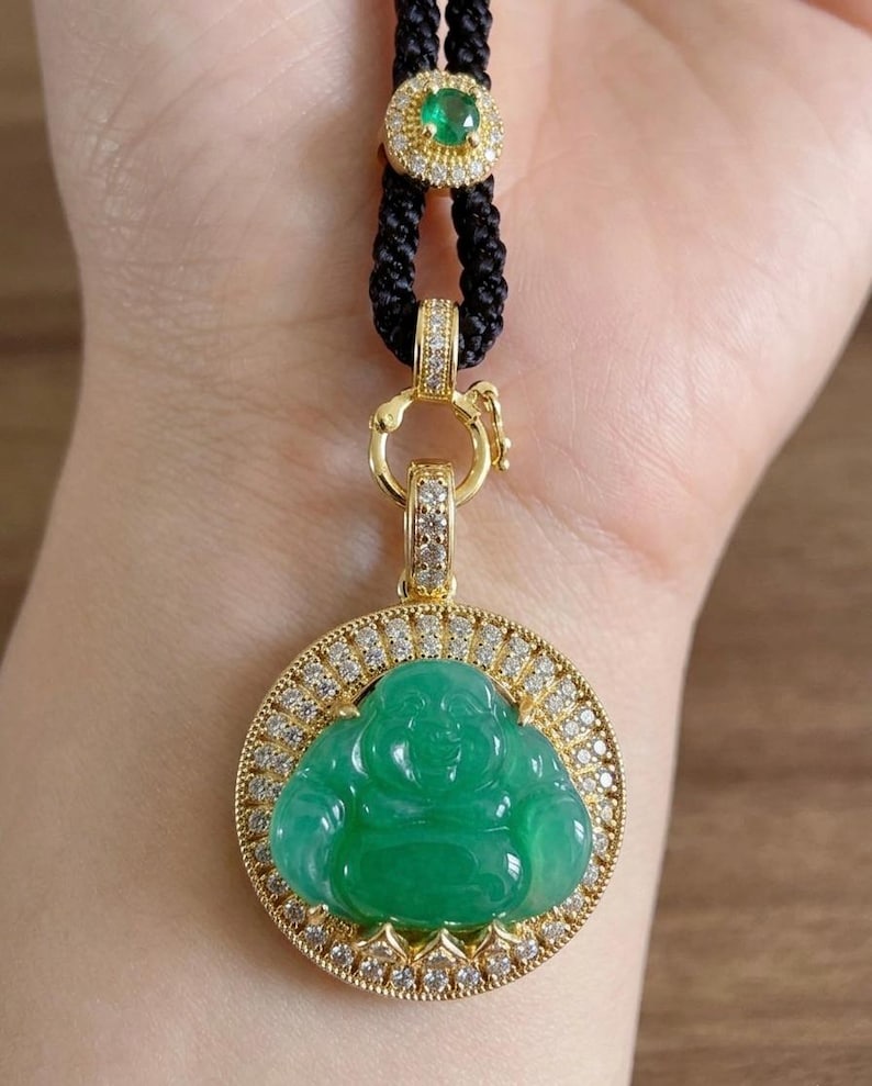 May include: A circular pendant featuring a carved green jade Buddha figure set in a gold frame encrusted with small white diamonds. The pendant hangs from a black braided cord adorned with a small gold accent piece containing a green stone.