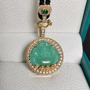 May include: A gold pendant featuring a carved green jade-like Buddha figure surrounded by a circular frame of small clear stones. The pendant hangs from a black cord with a gold accent piece containing a small green stone and clear stones.
