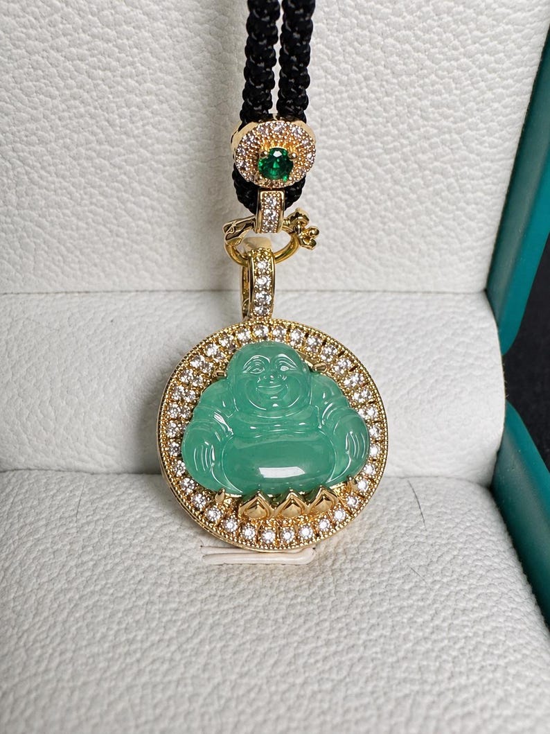 May include: A gold pendant featuring a carved green jade-like Buddha figure surrounded by a circular frame of small clear stones. The pendant hangs from a black cord with a gold accent piece containing a small green stone and clear stones.