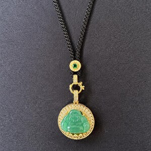 May include: A gold-toned necklace featuring a circular pendant with a green Buddha figure surrounded by small clear stones. The pendant hangs from a black textured cord accented with a small gold-toned bead containing a single green stone.