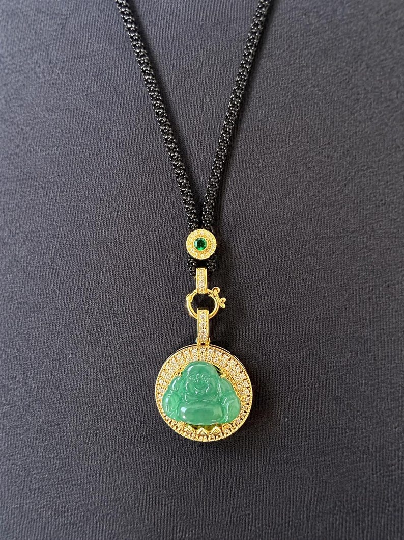 May include: A gold-toned necklace featuring a circular pendant with a green Buddha figure surrounded by small clear stones. The pendant hangs from a black textured cord accented with a small gold-toned bead containing a single green stone.