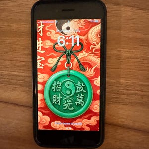 Op de afbeelding: Een zwarte smartphone met een vergrendelscherm met een rode en gouden achtergrond, Chinese karakters en een groene cirkelvormige hanger met een yin-yang symbool. De tijd is 6:11 uur en de datum is maandag 7 juli.