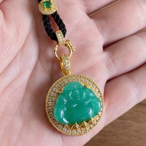 May include: A gold pendant featuring a carved green jade Buddha figure set within a circular frame encrusted with small white diamonds. The pendant hangs from a black braided cord accented with a small gold-rimmed green gemstone.