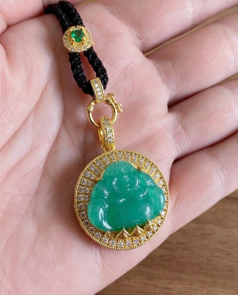 May include: A gold pendant featuring a carved green jade Buddha figure set within a circular frame encrusted with small white diamonds. The pendant hangs from a black braided cord accented with a small gold-rimmed green gemstone.