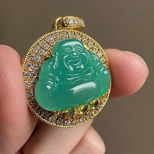 May include: A circular gold pendant featuring a central green jade-like figure of a laughing Buddha. The pendant is framed by a border of small, clear, sparkling stones and includes a gold bail at the top also set with clear stones.