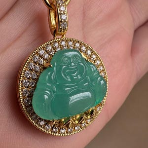May include: A circular gold pendant featuring a central carved green jade-like Buddha figure. The pendant is framed by a border of small, clear, sparkling stones and includes a gold bail also encrusted with clear stones for a necklace chain.