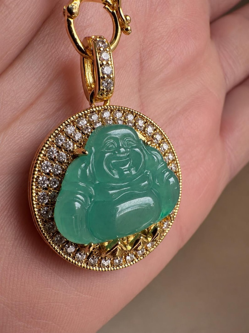 May include: A circular gold pendant featuring a central carved green jade-like Buddha figure. The pendant is framed by a border of small, clear, sparkling stones and includes a gold bail also encrusted with clear stones for a necklace chain.