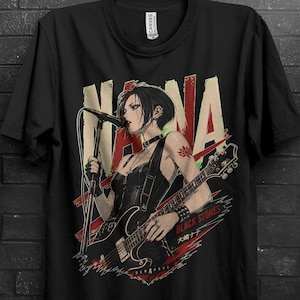 May include: A black short-sleeve t-shirt featuring a graphic print of a person with short dark hair singing into a microphone while playing an electric guitar. The design includes the text NANA, BLACK STONES, and Japanese characters in red and white.