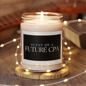 Könnte beinhalten: Eine weiße Kerze mit einem schwarzen Etikett, auf dem "Scent of a Future CPA" steht. Die Kerze ist angezündet und steht auf einem Holzuntersetzer.
