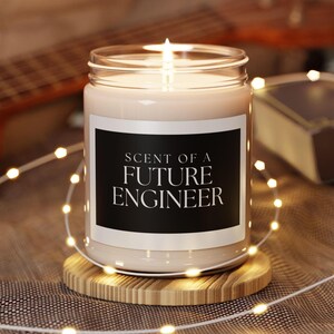 Könnte beinhalten: Eine weiße Kerze mit einem schwarzen Etikett, auf dem "Scent of a Future Engineer" steht. Die Kerze ist angezündet und steht auf einem Holzuntersetzer, um den weiße Lichterketten gewickelt sind.