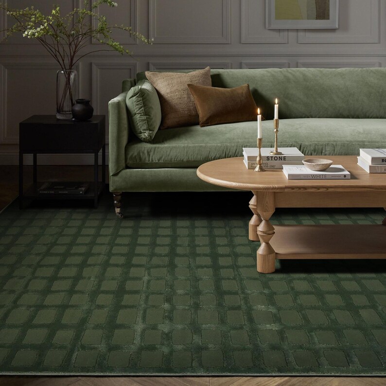 Op de afbeelding: Een groene vloerkleed met een rasterpatroon is het middelpunt van een woonkamer. Een groene fluwelen bank met bruine en beige kussens staat tegen een grijze muur. Een houten salontafel en een zwart bijzettafeltje maken de sc&egrave;ne compleet.