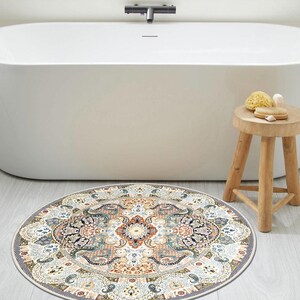 Puede incluir: Una alfombra de ba&ntilde;o redonda y estampada con un dise&ntilde;o detallado y ornamentado en tonos crema, azul, naranja y marr&oacute;n. La alfombra est&aacute; colocada frente a una ba&ntilde;era blanca, con un peque&ntilde;o taburete de madera y accesorios de ba&ntilde;o cerca.