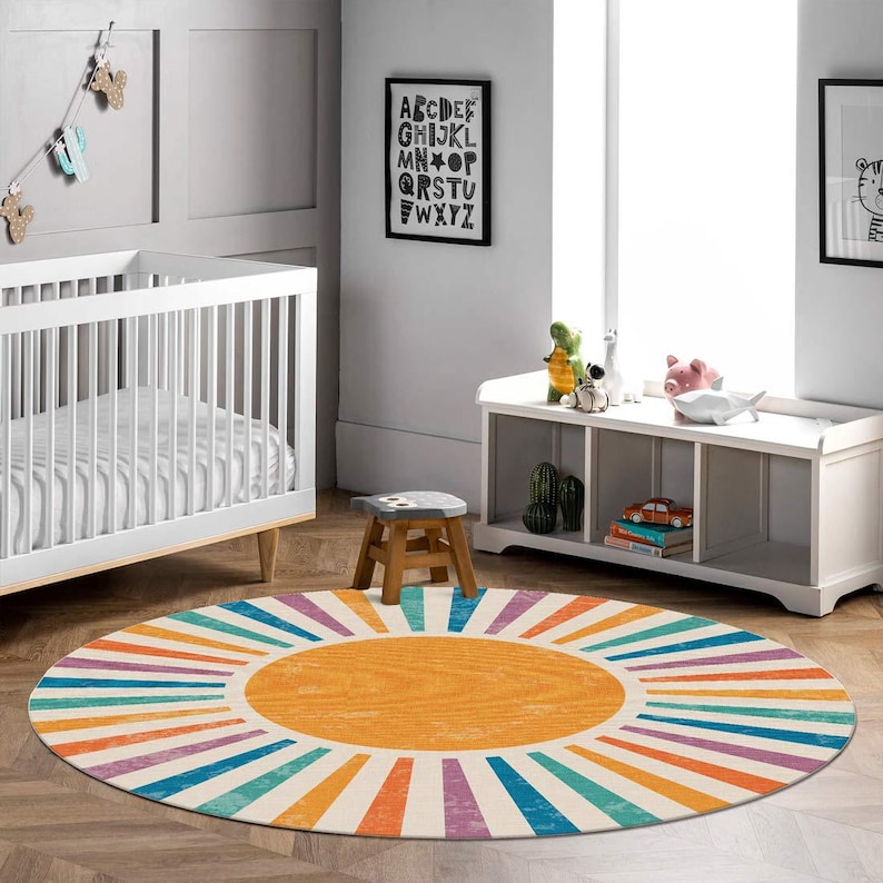 Op de afbeelding: Een kleurrijk rond tapijt met een zonnestralenontwerp in tinten oranje, geel, blauw, groen en paars. Het tapijt heeft een witte achtergrond met een grote oranje cirkel in het midden.