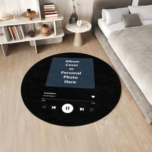 Pode incluir: Um tapete redondo preto com uma superfície texturizada, apresentando um design de interface de reprodutor de música. Inclui um espaço reservado para uma capa de álbum ou foto, juntamente com controles de música e texto. O tapete está sobre um chão de madeira.