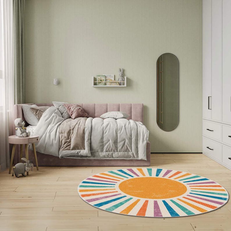 Op de afbeelding: Een roze bed met witte beddengoed en een kleurrijk rond tapijt met een zonnestralenontwerp. Het tapijt heeft oranje, gele, blauwe, groene en paarse strepen.
