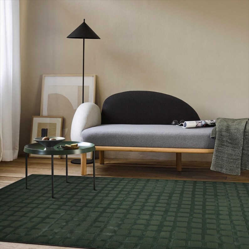 Op de afbeelding: Een moderne woonkamer met een donkergroen patroonkleed, een grijze bank met een zwarte rugleuning en een kleine groene bijzettafel. Een zwarte vloerlamp en ingelijste kunstwerken maken de inrichting compleet. De kamer heeft een neutraal kleurenpalet.
