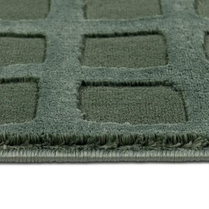 Op de afbeelding: Close-up van een getextureerd, saliegroen tapijt met een geometrisch vierkant patroon. Het tapijt heeft een zachte, zachte uitstraling met een donkerder groen verzonken ontwerp. De randen van het tapijt zijn netjes afgewerkt.