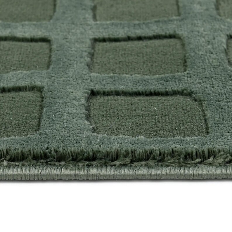 Op de afbeelding: Close-up van een getextureerd, saliegroen tapijt met een geometrisch vierkant patroon. Het tapijt heeft een zachte, zachte uitstraling met een donkerder groen verzonken ontwerp. De randen van het tapijt zijn netjes afgewerkt.