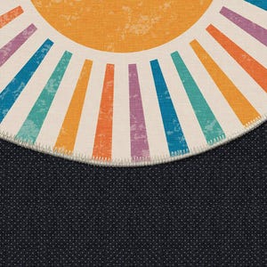 Op de afbeelding: Een rond tapijt met een witte achtergrond en een kleurrijk zonnestralenontwerp. De zonnestralen zijn gemaakt van oranje, gele, blauwe, groene en paarse strepen. Het tapijt heeft een zwarte, textuurrand.