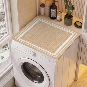 May include: A rectangular beige and white woven rug sits atop a white washing machine. The rug has a square pattern with a white border. The washing machine is in a well-lit laundry room with various toiletries on a shelf.