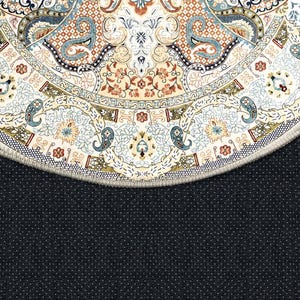 Puede incluir: Primer plano de una alfombra redonda con un intrincado dise&ntilde;o floral y paisley. La alfombra presenta un fondo crema con patrones en tonos azules, marrones y beige. El borde de la alfombra es visible, con un reverso azul oscuro punteado.