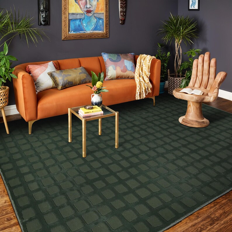 Op de afbeelding: Een donkergroen vloerkleed met een rasterpatroon bedekt een houten vloer. Een oranje bank met sierkussens en een kleine glazen tafel met gouden poten staan op de achtergrond. Een houten stoel in de vorm van een hand en potplanten maken de kamer compleet.