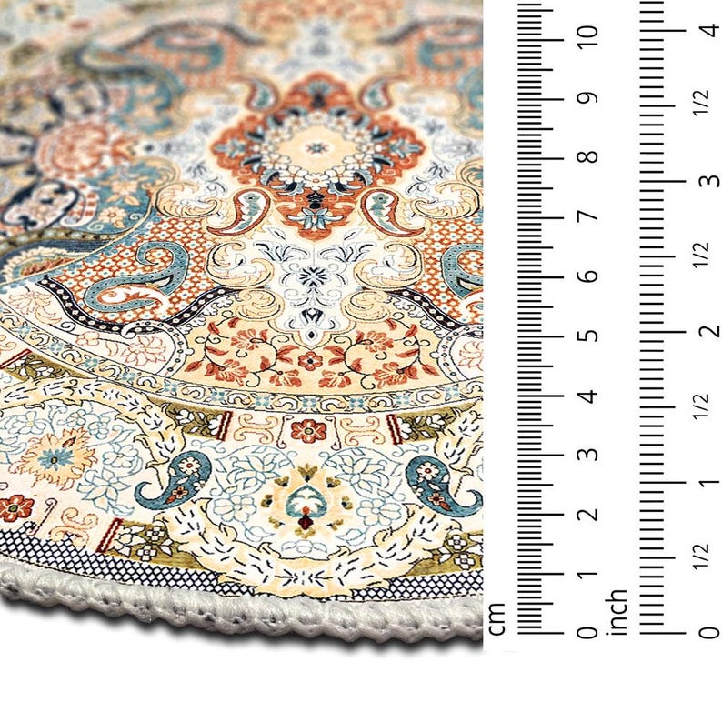 Puede incluir: Primer plano de una alfombra redonda con un patr&oacute;n intrincado y flecos color crema. La alfombra presenta un dise&ntilde;o floral detallado en tonos beige, naranja y azul. La regla muestra una medida de 10 cm.