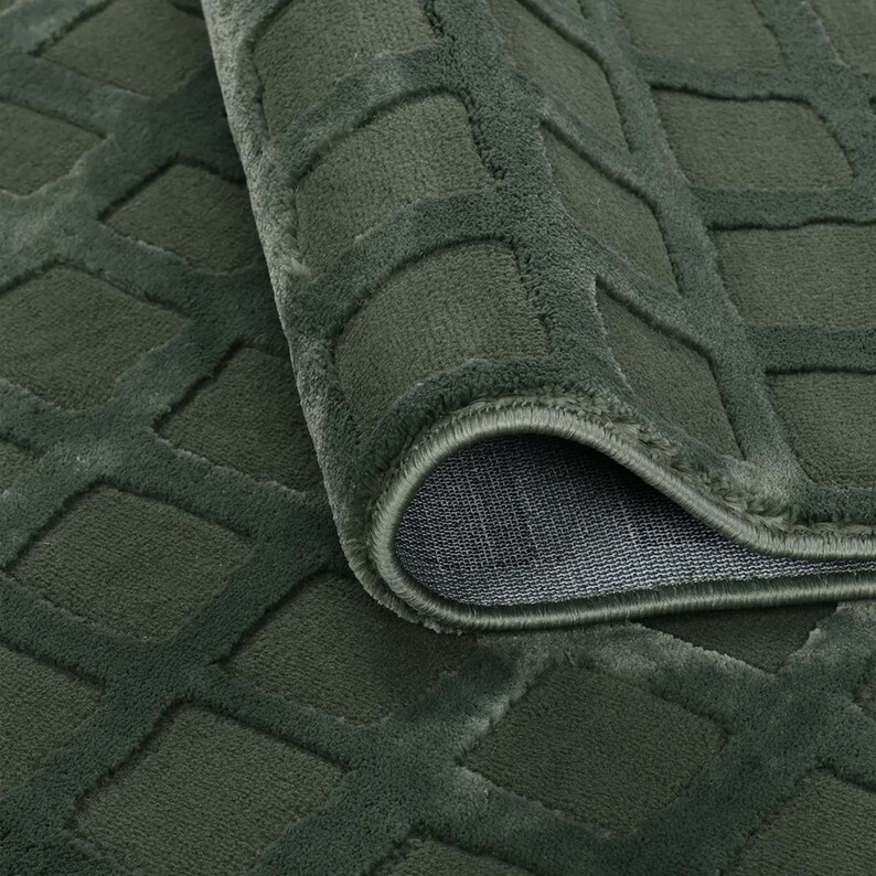 Op de afbeelding: Close-up van een opgerolde, olijfgroene vloerkleed met een geometrisch ruitpatroon. De textuur van het vloerkleed is zacht en het ontwerp is licht verhoogd, waardoor een driedimensionaal effect ontstaat. De onderkant van het vloerkleed is zichtbaar.
