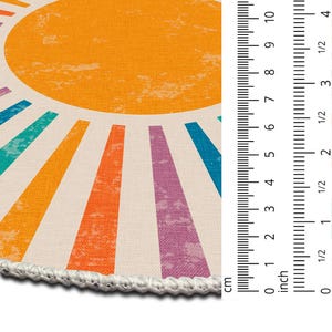 Op de afbeelding: Een rond, wit en oranje kleed met een zonontwerp. De zon is geel met oranje stralen. De stralen zijn omrand met verschillende kleuren, waaronder blauw, groen, paars en oranje.